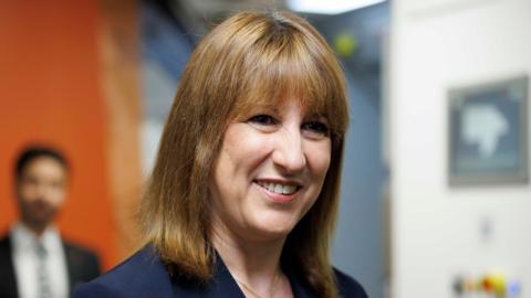 A woman with auburn shoulder-length hair is smiling. She is wearing a dark navy suit jacket and there is a man blurred in the background.