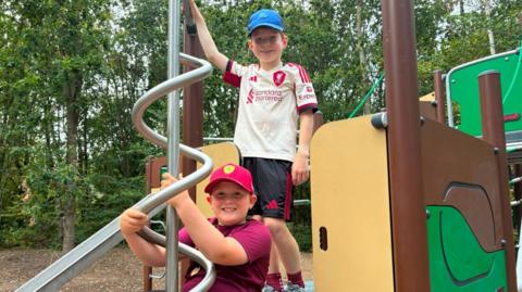 Two young boys wearing T-shirts, shorts and baseball caps are smiling while playing on climbing equipment at the park.
