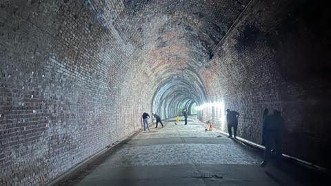 A long brick tunnel with people in the distance sweeping the ground. 