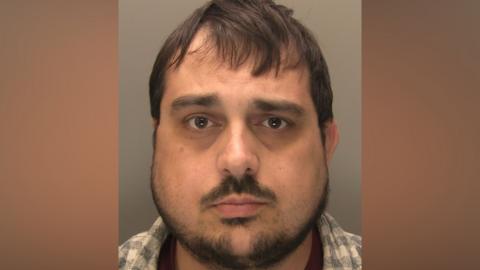 Police mug shot of a man with dark hair and a beard. He is wearing a checked shirt and looking directly at the camera.