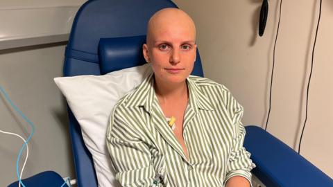 A young girl with no hair sits in a hospital chair looks into the camera, she's wearing a green and white stripped shirt. 