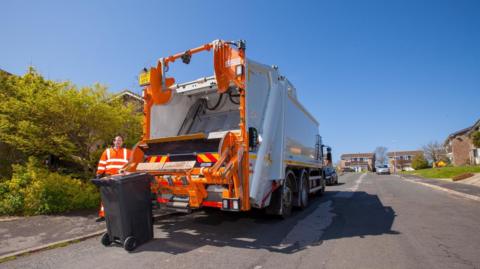 A white and orange bin collection lorry stopped at the side of a residential street. A man in orange overalls stands at the back with a black wheelie bin. It's a sunny day and the sky is blue.