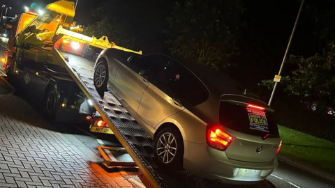 An estate car is hauled onto a tow truck after being seized by police. It is dark and the car has a sticker on its back window saying "seized by police".