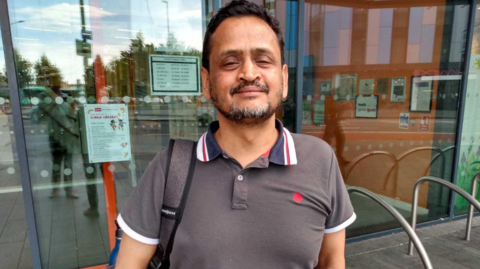 A man with a goatee and a moustache, wearing a brown polo shirt and carrying a rucksack on one shoulder, smiles at the camera. Behind him is a large set of glass doors.