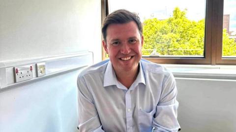 A man with short brown hair. He is sat down. He is wearing a light blue shirt and is smiling. Behind him is a white wall and a window. 