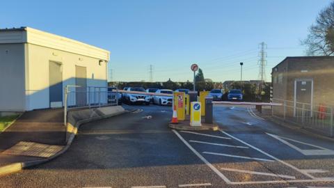 Cars parked in a car park in front of a hedge and behind an entrance with barriers across the entrance and exit lanes. A small brick building is on the right of the entrance with a white building on the left.