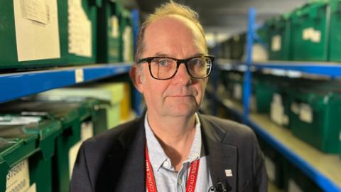A man in dark glasses is standing in an archive with blue metal shelves behind him on which there are green plastic boxes. He is wearing a dark blazer with a light grey shirt underneath and a red Bristol City Council lanyard.