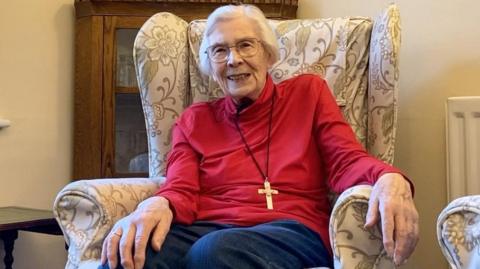 A woman with white hair, glasses and wearing a red top smiles into the camera. She has a wooden cross around her neck.