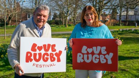 Councillors Neil Sandison and Maggie O'Rourke stand in a park holding "Love Rugby" boards. It's a sunny day and there are trees and flowers around them.