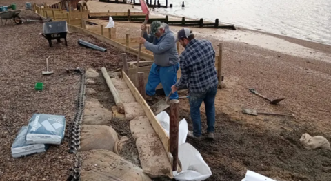 Two men trying to install some kind of barrier in the beach to halt the impact of coastal erosion. They are holding spades, there is a wheelbarrow, and bags of some type of grit. The sea is also in shot.