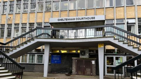The entrance of Cheltenham Magistrates' Court. It is a 1960's building and the entrance in on the first floor, accessed by two flights of stairs on either side. Above the entrance doors is a sign that reads "Cheltenham Courthouse".