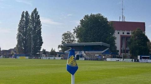 The picture shows a corner flag in the foreground, with a playing field and a stand in the background.