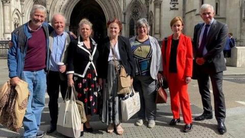 Four men and three women outside the stone-built Royal Courts of Justice in the Strand, London. They are carrying bags and rucksacks and standing in a line with the entrance to the court in the background. One is wearing a red jacket and trousers.