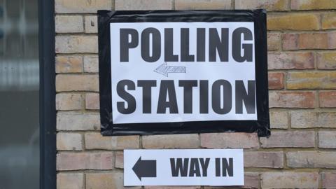 A generic sign stuck on a brick wall that reads polling station. Another sign below it details the way inside the polling station to the left. 