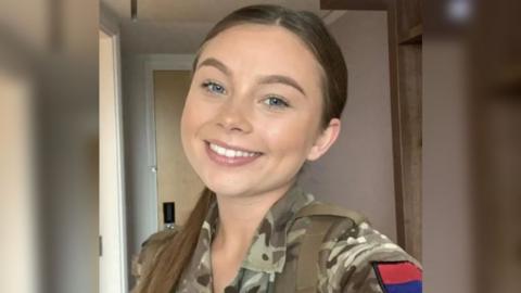 A selfie of Jaysley Beck wearing her military uniform, with her long brown hair slicked back into a low ponytail. She has blue eyes and is smiling at the camera.