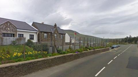 Three stone buildings in a line to the left with grass and tarmac in front of them. To the right is a straight main road. It is a cloudy day.