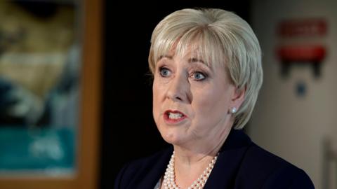 Heather Humphreys, a woman with short blonde hair, pictured from the shoulders up. She is wearing a black blazer, a pearl necklace and silver earrings. The background of the image is blurred. 