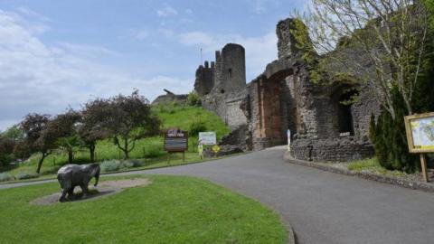 The outside of the castle grounds showing part of the ruined fortification with a large arched gateway. There is a small elephant statue on the green lawn in front, a small green hillside leading up to part of the ruins, some trees and signage. 