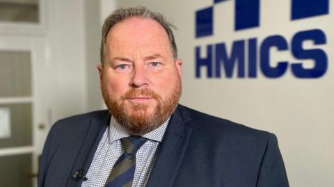 A man with short greying hair and a ginger beard looks at the camera. He is wearing a dark blue suit, chequered shirt and blue and green tie. He is standing in front of a blue HMICS sign. 