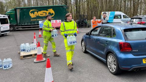 Two workers in hi-viz jackets carrying multi-packs of water towards a car.