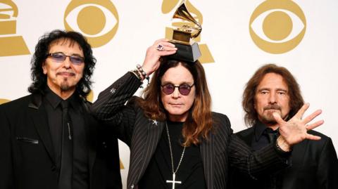 Bandmates Tony Iommi, Ozzy Osbourne and Geezer Butler pose in front of yellow and white Grammy logos while holding a grammy award for Best Metal Performance at the 2014 Grammy awards
