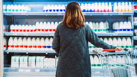 A female shopper, pictured from behind, with longish brown hair and a quilted navy long coat, stares at a fridge in a supermarket full of different types of milk. The brand is not shown. 