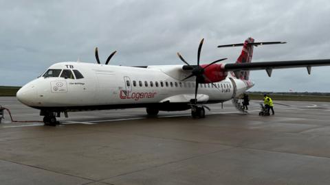 A Loganair aircraft on a tarmac. There is an airport worker in high-visibility clothing attending to the rear door.