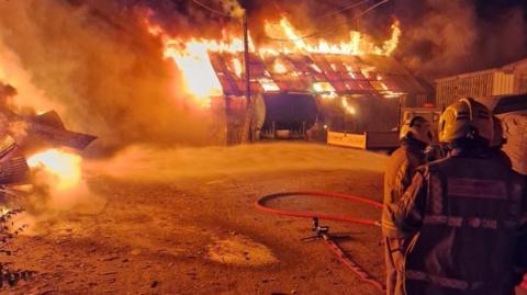 The backs of three firefighters can be seen in the foreground and a large buildng is on fire in the background.