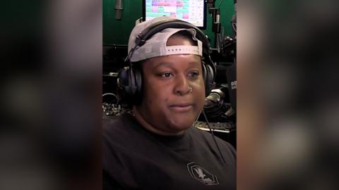 Remi wears a cap backwards and a black tshirt and has headphones on