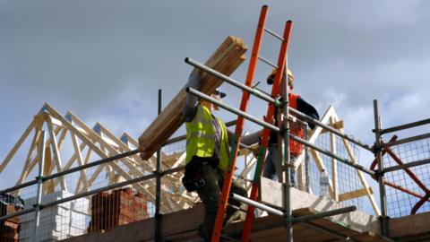 A photo of two builders on scaffolding. One is wearing a yellow hi-vis vest and is carrying planks of wood while climbing up an orange ladder. The other is wearing a hi-vis orange vest and is standing on scaffolding near the top of the ladder.