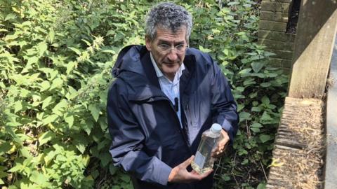 Peter Prinsley wearing a dark blue jacket and glasses, he has greying hair and is holding a plastic bottle of water. 

He is stood in what appears to be a stream with green foliage around him and a bridge to the right of the image. 