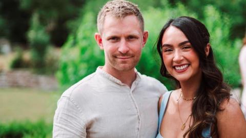 Joe and Jessie smiling into the camera. Joe is standing on the left and has blonde, short hair with stubble around his mouth. He is wearing a cream shirt and is smiling with his mouth closed. Jessie is on the right and has long, dark hair which has been curled. She is smiling with her teeth and has red lipstick on. She is wearing two gold necklaces and a blue square-neck top with wide straps. They are in a garden, but the background is out of focus.