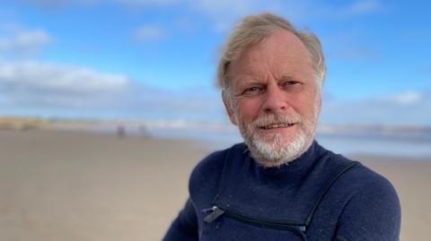 Kitesurfer Adrian Cooke on the beach at Fraisthorpe, with the blue sea and sky in the background. He has light brown hair and a white beard, and wears a dark blue wetsuit.