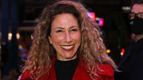 A woman with long, curly brown hair smiles at the camera. She wears red lipstick, a red coat and a black top.