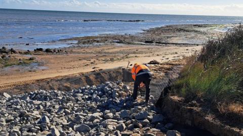 A worker at the top of the beach at Walton-on-the-Naze. He is standing on large stones looking towards the sea