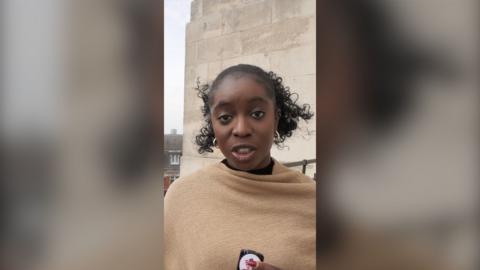 A woman with curly black hair is wear a camel-coloured wrap. She is speaking into the camera and holding a button mic.