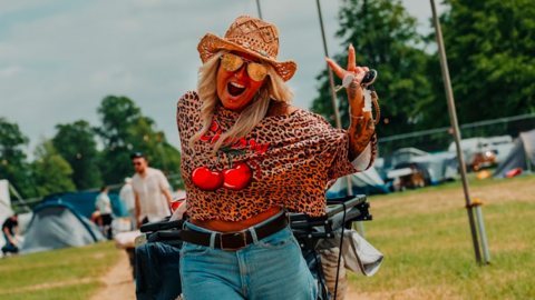 Woman in a top with cherries on it. She smiles widely and walks through a campsite while pulling luggage behind her.