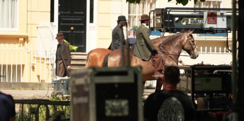 Two men in traditional clothing riding brown horses near houses.