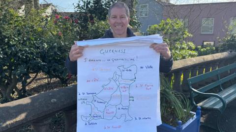 Man standing in a park in front of a bench. He is holding a map with the parishes in Guernsey written in Guernésiais.