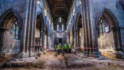 A group of people in high vis vests are standing in the centre of St Cuthbert's in Darlington with the floor removed exposing old walls beneath the church. 