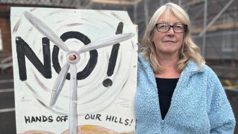 A woman in a light blue fleece top and black T-shirt with long blonde hair and glasses holds up a "Hands Off Our Hills" placard