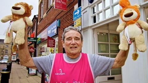 A man wearing a pink running bib with a grey t-shirt underneath is standing on a street in front of a shop and holding up two soft toy lions