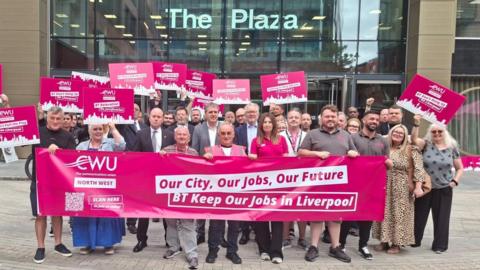 A large group of people are standing outside an office building called The Plaza. They are holding pink placards which say "BT don't pull the plug" and other slogans. The people at the front are holding a large banner which says "BT keep our jobs in Liverpool".