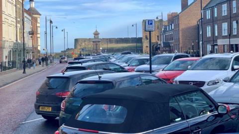 A view down Tynemouth Front Street, with cars parked diagonally in spaces down the middle of the road. The street is lined with three-storey Georgian buildings, some with shop fronts, and ends with a view of a clock tower and old castle walls.