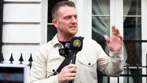 Tommy Robinson, standing outside Westminster Magistrates' Court, in London, talking into a black microphone, with a golden lion head on it, with his left hand raised. He is wearing a light jacket, a black T-shirt and has short dark hair and short beard. He is standing by black railings. 
