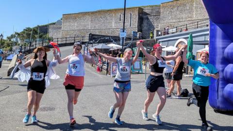 The finish line of a pirate fun run. There are five women running under a blue inflatable, they are wearing numbers pinned to their tops and wearing shorts, tank tops and trainers. The woman on the left is wearing a pirate hat and the other women are wearing bandanas. 