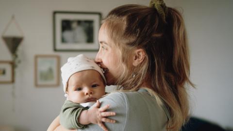 A mother smiles as she holds her baby. The baby has a white hat on and a hand outstretched. The mother is not looking at the camera. The way her face is lit up suggests she is looking out of a window.