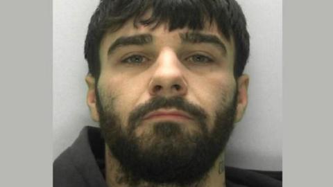 Connor Parsons looks into the camera for his police custody image. He has black hair, a thick beard and wears what looks like a black hoodie.