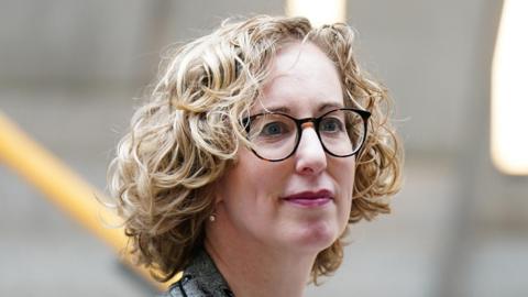 Lorna Slater has blonde, curly hair and is wearing glasses and a grey jacket
