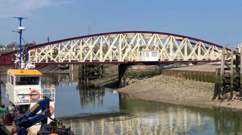 Ramsey Swing Bridge, a curved red and white structure with white x-shaped metal bars. The water is flat calm and there is a fishing boat in the foreground to the left.